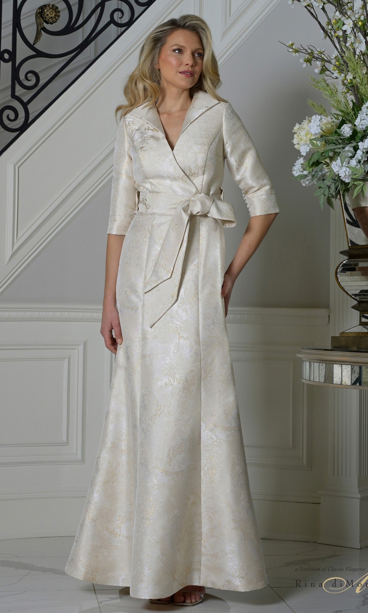 A woman stands indoors near a staircase and floral arrangement wearing the Rina Di Montella RD3521 long formal dress in champagne, featuring jacquard fabric, three-quarter sleeves, and a sash belt.